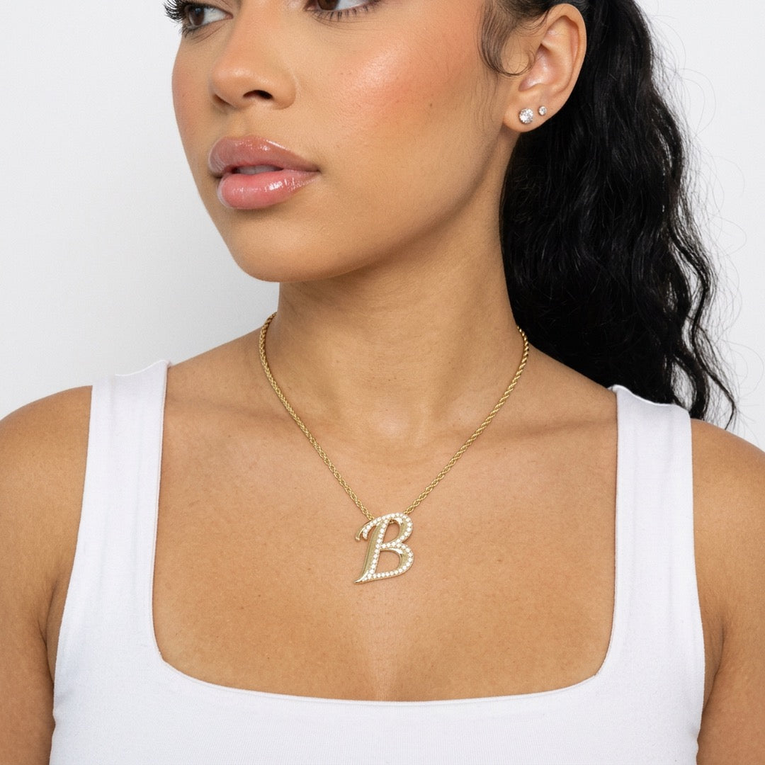 A woman wearing a delicate gold chain necklace with a capital letter 'B' pendant paved with small clear stones, paired with a white tank top.